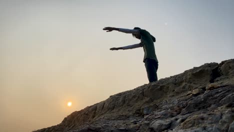 Young-man-exercises-outdoor-on-top-of-rocky-hill,-sunset,-static,-slowmo
