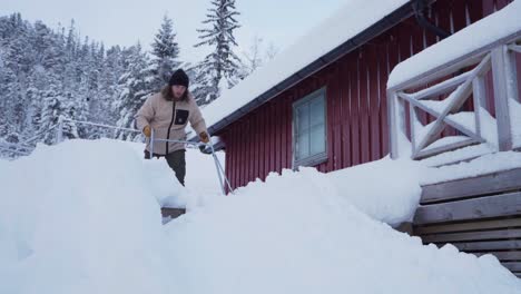 Man-Cleaning-Scraping-Snow-Off-The-Cabin-Using-Sleigh-Shovel-In-Indre-Fosen,-Norway---low-angle