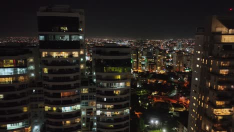 Aerial-trucking-shot-of-modern-luxury-high-rise-apartments-and-beautiful-illuminated-city-of-Santo-Domingo-in-background-at-night