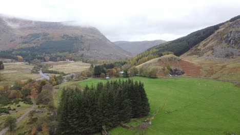 Aerial-view-between-the-green-hills-and-the-river-of-the-Scottish-highlands-with-trees-and-yellow-leaves,-misty-and-cloudy