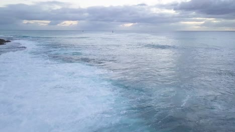 Hellblaue-Wellen-Und-Weißer-Meeresschaum,-Der-Auf-Eine-Felsige-Küste-In-West-Oahu-Honolulu-Koolina-Hawaii-Mit-Segelbooten-Im-Sonnenuntergangshintergrund,-Luftwagen