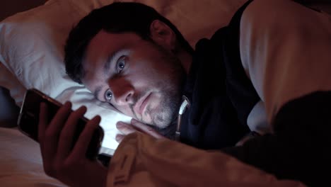 Man-lying-in-bed-with-smartphone,-screen-lights-up-face-in-dark-bedroom,-scrolling,-social-media,-texting-friends-in-night-before-sleep
