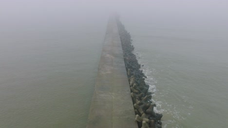 Aerial-establishing-view-of-Port-of-Liepaja-concrete-pier,-Baltic-sea-coastline-,-foggy-day-with-dense-mist,-moody-feeling,-big-storm-waves-splashing,-wide-birdseye-drone-shot-moving-forward