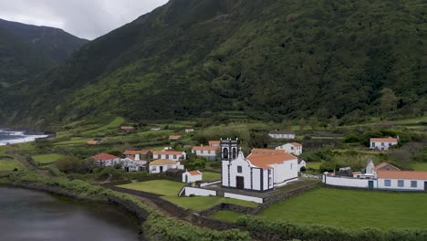 Drone-orbiting-view-of-a-coastal-village-church,-lush-green-cliffs-landscape,-Fajã-de-Santo-Cristo,-São-Jorge-island,-the-Azores,-Portugal