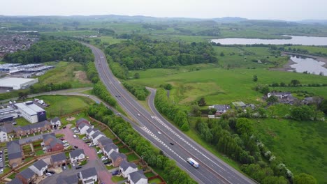 Aerial-video-of-Scottish-Moterway-Countryside,-freeway-with-traffic-surrounded-by-green-vegetation-and-buildings-on-one-side-and-lakes-of-water