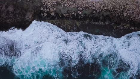 Top-down-aerial-waves-crashing-over-a-rocky-shoreline-with-beautiful-white-sea-foam-and-vibrant-blues-in-oahu-hawaii-honolulu,-AERIAL-DOLLY-TOP-DOWN