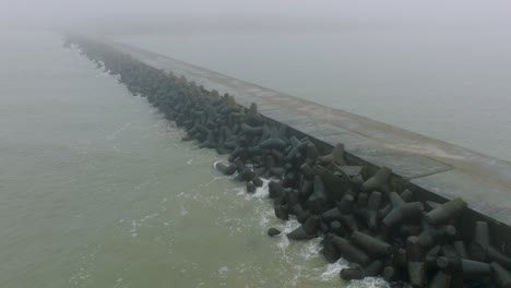 Aerial-establishing-view-of-Port-of-Liepaja-concrete-pier,-Baltic-sea-coastline-,-foggy-day-with-dense-mist,-moody-feeling,-big-storm-waves-splashing,-wide-drone-dolly-shot-moving-left