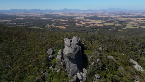 Aerial-of-man-made-lookout-atop-giant-boulder-in-Australian-bush,-mountains-in-background