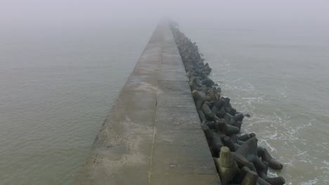 Aerial-establishing-view-of-Port-of-Liepaja-concrete-pier,-Baltic-sea-coastline-,-foggy-day-with-dense-mist,-moody-feeling,-big-storm-waves-splashing,-birdseye-drone-shot-moving-forward-low
