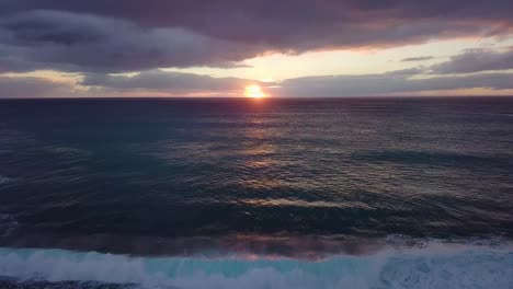 Beautiful-sunset-over-west-oahu-honolulu-hawaii-with-golden-hour-light-reflecting-off-the-ocean-and-crashing-white-cap-waves-on-a-rocky-sea-shore,-AERIAL-TILT-DOWN