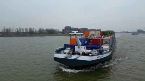 Aerial-Dolly-Around-Forward-Bow-Of-Scaldis-Container-Ship-Travelling-Along-River-On-Overcast-Day-In-Dordrecht