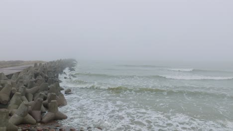 Aerial-establishing-view-of-Port-of-Liepaja-concrete-pier,-Baltic-sea-coastline-,-foggy-day-with-dense-mist,-moody-feeling,-big-storm-waves-splashing,-wide-drone-shot-moving-forward-low