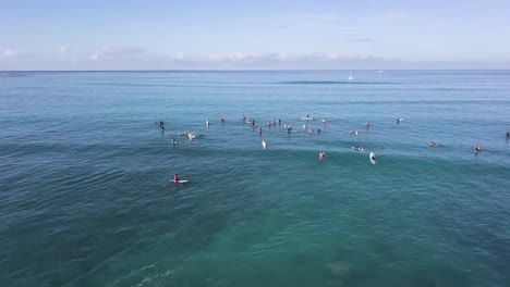 Crowd-of-surfers-at-waikiki-beach-honolulu-hawaii-with-a-pan-reveal-of-mountains-and-honolulu-city,-AERIAL-TRUCKING-PAN