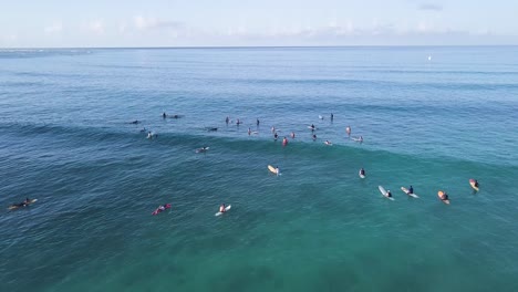 Slow-movement-over-a-large-group-of-surfers-on-top-of-crystal-clear-blue-water-at-waikiki-beach-in-honolulu-hawaii,-AERIAL-DOLLY-TILT