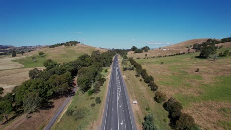 Cars-driving-on-a-straight-country-road-with-rolling-green-hills,-aerial