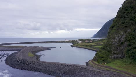 Drone-view-reveiling-a-coastal-village,-lush-green-cliffs-landscape,-lagoon,-Fajã-de-Santo-Cristo,-São-Jorge-island,-the-Azores,-Portugal