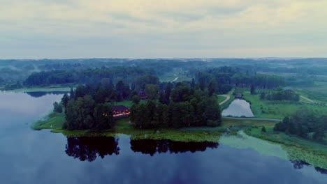 Beautiful-aerial-of-a-house-surrounded-by-trees-on-a-half-island-on-a-cloudy-day-with-everything-being-mirrored-in-the-still-lake