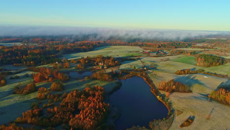 Stunning-aerial-of-lake,-frosty-fields-and-autumn-trees-in-the-sunlight-with-blue-skies-and-fog-moving-in-the-horizon
