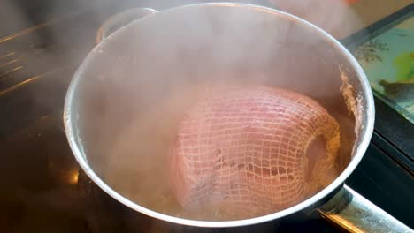 Cured-gammon-hind-leg-of-pork-in-netting-simmering-and-cooking-in-a-saucepan-in-the-kitchen-for-Sunday-roast-dinner