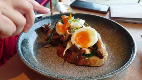 Person-eating-perfectly-cooked-poached-eggs-on-sourdough-bread-with-mushrooms-for-breakfast-in-a-local-cafe,-close-up-of-delicious-brunch-of-eggs-on-toast