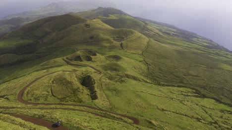 drone-dolly-view-of-vulcanic-mountains-covered-in-lush-green-vegetation-with-dirt-road,-cloudy-weather-in-São-Jorge-island,-the-Azores,-Portugal