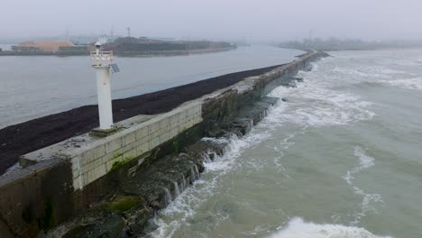 Aerial-establishing-view-of-Port-of-Liepaja-concrete-pier,-Baltic-sea-coastline-,-foggy-day-with-mist,-moody-feeling,-big-storm-waves-splashing,-white-lighthouse,-drone-shot-moving-forward