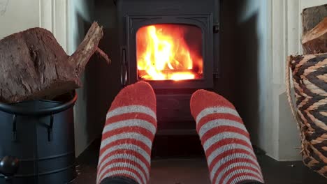 Warming-up-cold-feet-wearing-striped-wool-hiking-socks-in-front-of-log-burner-fire-during-chilly-winters-day-in-cosy,-warm-house