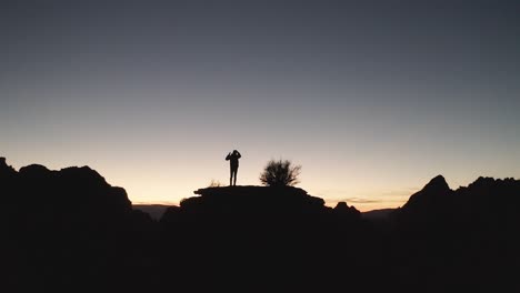 Silhouette-of-a-man-on-a-hill,-contemplating-life-as-he-looks-out-of-a-vast-desert-sunset
