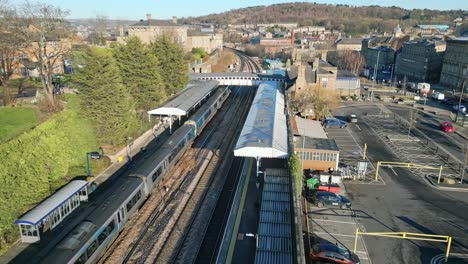 Cinematic-aerial-drone-footage-of-train-leaving-the-train-station-with-platform-and-carpark-Dewsbury-West-Yorkshire,-UK