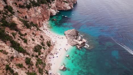 Aerial-view-of-white-sand-beach-on-rocky-coastline-with-turquoise-water,-Sardinia