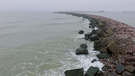 Aerial-establishing-view-of-Port-of-Liepaja-stone-pier,-Baltic-sea-coastline-,-foggy-day-with-dense-mist,-moody-feeling,-big-storm-waves-splashing,-wide-drone-shot-moving-forward-low