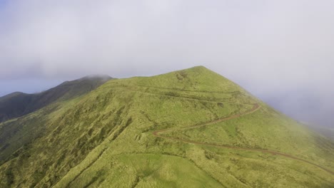 Drone-orbiting-vulcanic-mountain,-Pico-da-Esperança,-covered-in-lush-green-with-low-clouds-in-São-Jorge-island,-the-Azores,-Portugal