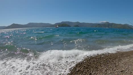Wide-view-of-boats-and-small-waves-lapping-onto-stony-shore-in-summertime---Karaka-Point,-Queen-Charlotte-Sound