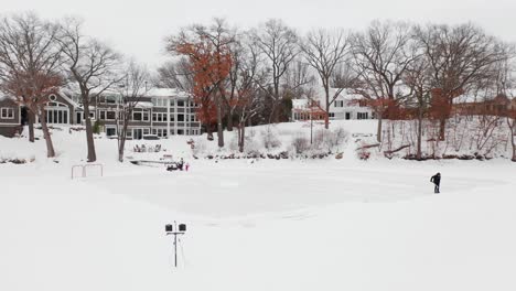 Aerial,-person-shoveling-snow-to-create-a-small-ice-hockey-rink-on-backyard-pond