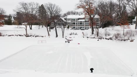 Aerial,-person-clearing-snow-to-make-a-small-hockey-ice-rink-on-a-frozen-lake-outside-of-house