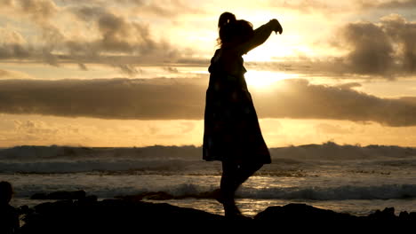 Free-spirited-girl-in-dress-swirls-around,-silhouette-against-gold-ocean-sunset
