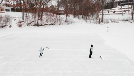 Aerial,-two-kids-clearing-snow-and-ice-skating-on-outdoor-ice-hockey-rink-on-a-frozen-lake