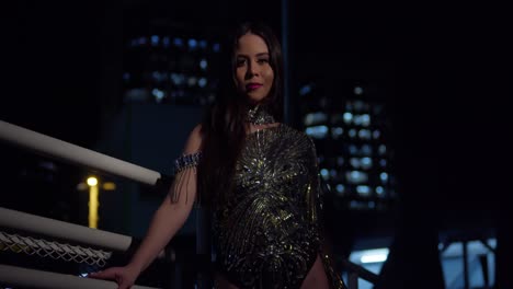 Young-latina-stands-on-a-staircase-at-night-in-a-sparkle-dress-with-city-lights-in-the-background