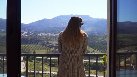 Caucasian-girl-with-straight-hair-and-a-checkered-coat-goes-through-a-window-and-walks-towards-a-sunny-background-of-mountains-and-fields