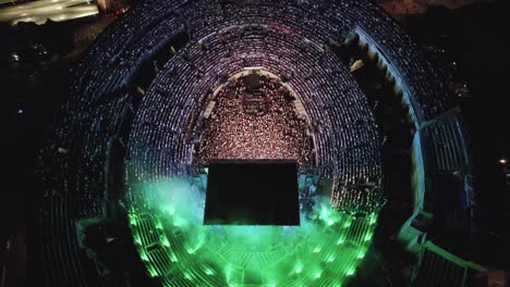 Drone-on-the-Arenas-de-Nîmes-in-the-middle-of-the-night,-people-are-watching-the-concert-and-there-are-lights-of-several-colors-and-city