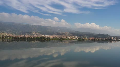 The-town-of-Paipa-and-its-surrounding-hills-is-reflected-in-lake-Sochagota,-captured-on-a-calm-day-with-a-4K-drone-slowly-moving-left-to-right-and-gaining-altitude