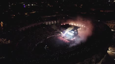 Drone-on-the-Arenas-de-Nîmes-in-the-middle-of-the-night,-people-are-watching-the-concert-and-there-are-lights-of-several-colors
