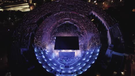 Aerial-view-above-the-Arenes-de-Nimes-at-night-during-a-show,-the-arenas-are-packed-with-people