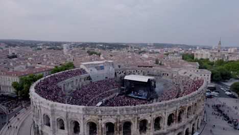 Drone-in-over-the-Arena-of-Nimes-at-sunset,-people-are-waiting-for-the-stromae-concert