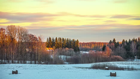 Zeitraffer-Einer-Verschneiten-Landschaft-Mit-Bäumen-Und-Holzhütten-Und-Mit-Orangefarbenen-Wolken,-Die-In-Einem-Goldenen-Himmel-Nach-Links-Ziehen