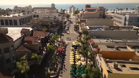 drone-video-of-Main-Street-in-Huntington-Beach,-California-leading-into-the-Pacific-Ocean,-showcasing-the-famous-pier-and-lively-beach-atmosphere-with-shopping-and-restaurants,-and-colorful-umbrellas