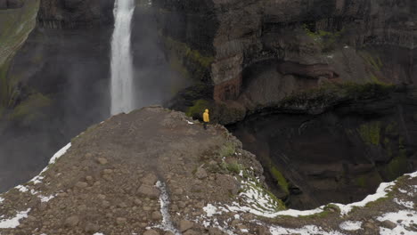 Aerial:-Slow-panning-shot-of-a-man-standing-near-the-edge-of-a-cliff,-watching-Haifoss-waterfall-in-Iceland