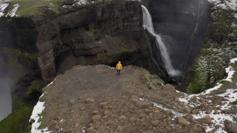 Aerial:-One-person-near-a-cliff,-wearing-yellow-jacket,-walks-towards-a-waterfall-in-Iceland