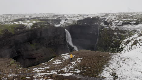 Aerial:-One-man-wearing-a-yellow-jacket,-walking-near-the-edge-of-a-cliff-towards-Granni-waterfall-in-Iceland