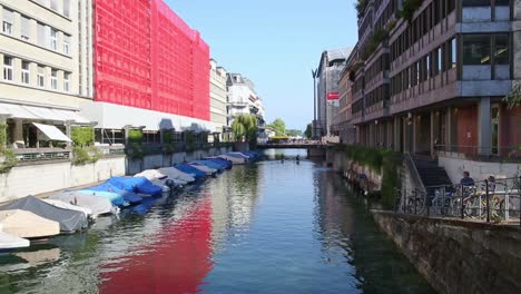 Kleine-Boote-Unter-Abdeckungen-Auf-Fluss-Und-Zürich-Stadt-Ruhige-Sonnendurchflutete-Straßenlandschaft